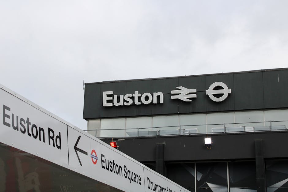A close-up view of the Euston underground station sign, featuring the station name 'Euston' in bold black letters, accompanied by the iconic London Underground and National Rail symbols in white. The sign is mounted on a black panel against a cloudy sky background. To the left, part of a white directional sign lists nearby locations including 'Euston Rd,' 'Euston Square,' and 'Drummond St,' with a black arrow pointing left. The scene captures the station's modern, clean signage structure with minimal lighting and a focus on clear, legible typography, providing a visual reference for public transport signage in London.