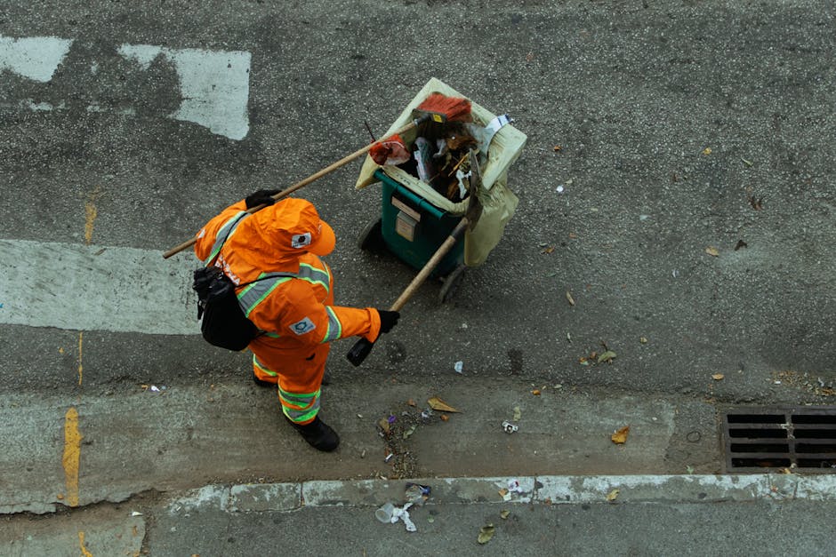 A worker from Kingston Cleaners wearing a high-visibility yellow vest and dark clothing operating a waste collection vehicle in an urban setting. The vehicle has a white body with red and orange refle
